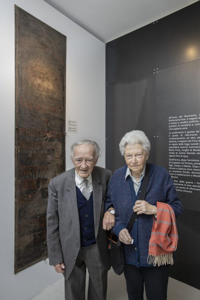 1. Cesare Finzi con la moglie Vera alla mostra del MEIS “Ferrara ebraica” vicino all’insegna della Profumeria Finzi donata al Museo.